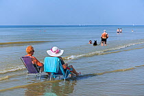 Two elderly ladies sunbathing in chairs on the beach just in the sea, and couple with dog paddling in water along the North Sea coast during heat wave, Belgium, July 2019