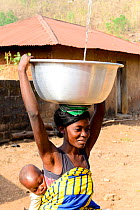 Woman carrying baby on back collecting water from fountain, dish balanced on her head. Dassa, Benin, 2020.