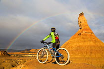 Cyclist beside Castildetierra mountain looking at rainbow over badlands of Bardenas Reales Natural Park. Navarre, Spain. February 2015.