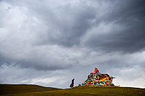 Woman approaching Buddhist chorten / stupa covered in prayer flags, under stormy sky. Ganden Thubchen Choekhorling Monastery, Litang, Garze Tibetan Autonomous Prefecture, Sichuan, China. October 2016....