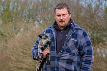 Young farmer holding Black welsh badger-faced mountain sheep lamb. Monmouthshire, Wales, UK, March