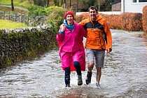 BBC environment correspondent Justin Rowlatt and woman wading along flooded road following Storm Ciara. Rothay Bridge, Ambleside, Lake District, UK. February 2020.