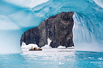 Tourists from expedition cruise ship in zodiac between ice arch and rock cliff on Spert Island. Palmer Archipelago, Antarctica. December 2019.