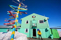 Man outside Vernadsky Station Ukrainian research base, signpost pointing to places across the world. Galindez Island, Argentine Islands, Antarctica. December 2019.