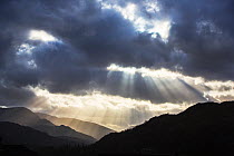 Sun rays shining through clouds onto Loughrigg Fell and hills of the Lake District National Park, in evening. Ambleside, Cumbria, England, UK. April 2020.