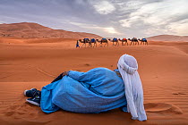 Berber man sits on the sand and watches as a camel caravan crosses the sane dunes of Erg Chebbi. Morocco.