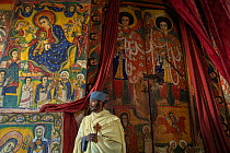 Young priest standing in front of mural paintings in the sanctum of Ura Kidane Mehret, an Ethiopian Orthodox Church. Zege Peninsula, Ethiopia. 2018.