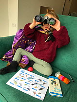 Young girl looking through binoculars, taking part in the RSPB Big Garden Birdwatch 2021; indoors looking out through window to feeders, with bird book, Bristol, UK. Model released. Medium repro only.