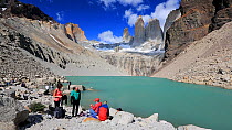 Panning shot showing people gathered near meltwater from retreating glacier flowing over rock into lake, below Torres del Paine towers. Torres del Paine National Park, Patagonia, Chile, January, 2020.