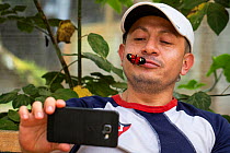 A man with a Heliconius butterfly on his face takes a selfie with his mobile phone, Mindo butterfly Farm (Mariposas de Mindo). Mindo, Ecuador.