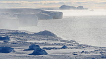 Sea fog moving across freshly formed sea ice, interrupted by open pools, ice shelf and iceberg in the distance, Antarctica, March.