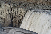 Dettifoss waterfall and tourists, in Vatnajokull National Park in Northeast Iceland. Eary spring run-off, with snow pack colored with lava dust. The falls drop 44 metres (144 ft).