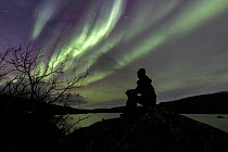 Man watching the Aurora borealis on a lakeshore, east of Abisko, Sweden. October.