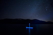Woman on a Stand up paddle board, lit with neon lights, on Loch an Eilian under a starry sky, Cairngorms National Park, Scotland, UK. March, 2021.