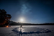 Woman ice skating on the frozen Loch Vaa at night beside a camp fire, Cairngorms National Park, Scotland, UK. January, 2021.