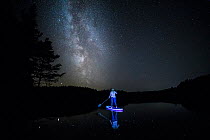 Woman on Stand up Paddleboard lit with neon lights on Uath Lochans at night, with the Milky Way across the night sky, Cairngorms National Park, Scotland, UK. September, 2020.