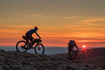 Two mountain bikers descending a mountain ridge at sunset, Cairngorms National Park, Scotland, UK. September, 2015.