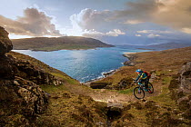 Mountain biker riding up rugged terrain with a view of surrounding islands, Isle of Harris, Outer Hebrides, Scotland, UK. April, 2015.