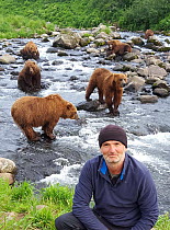 Portrait of photographer Andrew Parkinson in front of Brown bears (Ursus arctos beringianus) waiting for salmon to make way upstream, Kambalnaya River, Kamchatka Peninsula, Russian Federation. 2018