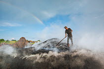 Man standing on pile of smouldering wood, making charcoal, with rainbow in background, Calonda, Harghita, Transylvania, Romania. June. Model released.