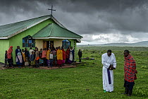 Local Maasai assistant Priest, Michael Chichilai, standing outside his church in the rain with his congregation after a Sunday service in Roman Catholic denomination church, Irkeepusi, Ngorongoro, Tan...