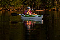 Woman in kayak on lake photographing wildlife in evening, New Hampshire, USA. July, 2020.