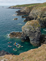 Group of sea kayakers approaching a natural rock arch, exploring rugged coastline near Porthclais, St. David's, Pembrokeshire, Wales, UK. August, 2022.