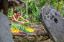 Young woman in traditional outfit for cultural ceremonies  weaving a basket from a palm frond whilst surrounded by stone moneys in a village, Yap, Micronesia.