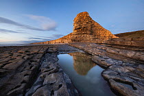 Tide pool in bedrock along the shore with towering cliffs in background at sunset, Nash Point, Glamorgan Heritage Coast, Wales, UK. December, 2022.