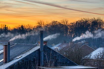 Domestic rooftop chimneys on houses emitting vapour from gas boilers for central heating at sunrise on freezing cold winter morning, Flanders, Belgium. December, 2021.