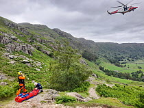 Two mountain rescue workers preparing to evacuate their casualty from a hillside to a waiting coast guard helicopter, Lake District, Cumbria, UK. June, 2022.