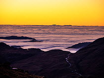 View from the Coniston fells over Harter Fell at sunset, shrouded in mist from a temperature inversion. Hardknott Pass road visible over the fells and Isle of Man in the distance on the horizon, Lake...