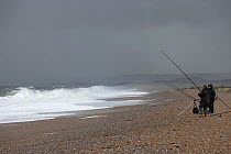 Two people fishing from the beach on a stormy day, Cley, Norfolk, UK. September, 2022.