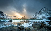 Winter storm clouds gathering along rocky shore surrounded by snow-covered mountains, Lofoten Island, Norway. March, 2023.