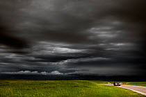 Car travelling along Badlands Loop Road under dark storm clouds, Badlands National Park, South Dakota, USA. June, 2022