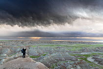 Photographer standing on rocky overlook at sunrise photographing rocky landscape and stormy sky, Big Badlands Overlook, Badlands National Park, South Dakota, USA. June, 2022.