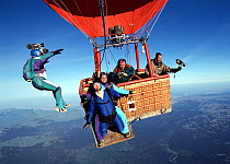 Base-jumper leaps off hot air balloon as two parachutists prepare to fall, a man films using a camcorder from the basket and another holds a trained Peregrine falcon (Falco peregrinus), Italy.