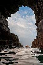 View looking out across sea from inside a sea cave, with a lifeboat approaching in background, St Tudwals Island East, Abersoch, Gwynedd, Wales, UK. July, 2016.
