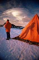 Man outside tent watches lenticular clouds forming over Coronation Island at sunset, South Orkneys, Antarctica.