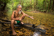 Village chief Apai Bandi sits on log on the side of his tribe's pristine river and talks to about the importance of protecting their forest, Utik River, Sungai Utik Customary Forest, Kalimantan,...