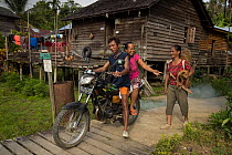 Iban man and woman on motorcycle, driving past house and woman carrying dog, Kapuas Ulu, Kalimantan, Borneo, Indonesia. All are heading out to work in fields.