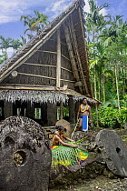 Man and woman in traditional, ceremonial dress outside a village hut. The young woman is weaving a basket from a palm frond and surrounded by stone money, Yap, Federated States of Micronesia. August,...