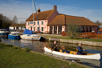 Two people in canoe paddling past boats moored along Hickling Broad with The Pleasure Boat public house in background, Norfolk Broads National Park,  Norfolk, UK. March, 2007.