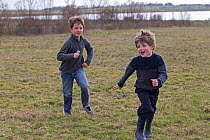 Boys chasing each other in field with river behind, Norfolk, England, UK. April, 2015. Model released.