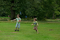 Two boys running through field and blowing bubbles with toy, Norfolk, England, UK. July, 2014. Model released.