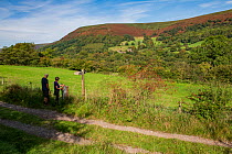 Hikers walking along footpath through Vale of Ewyas with Llanthony Priory and Hatterall Ridge behind, Brecon Beacons National Park, Monmouthshire, Wales, UK. October, 2023. Model released.