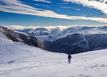 Cloud spilling over Glaramara Fell with a woman hiker descending snow-covered Robinson Fell in winter, Lake District, Cumbria, UK. January, 2023.