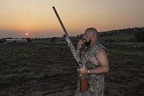 Maltese hunter walking through farmland with shotgun and blowing whistle to control hunting dog at dawn, whilst hunting Common quail (Coturnix coturnix), Siggiewi, Malta. September, 2020. Model releas...