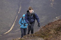 Boy and girl hiking up steep mountainside, above Achlean, Cairngorms, Scotland, UK. April, 2023. Model released.