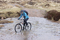 Girl on mountain bike cycling through water, north of Loch Eanaich, Cairngorms, Scotland, UK. April, 2023. Model released.